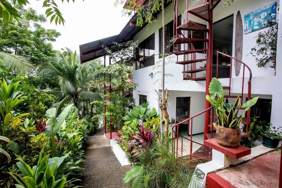 A red spiral staircase climbs the white building beside a garden walkway full of plants