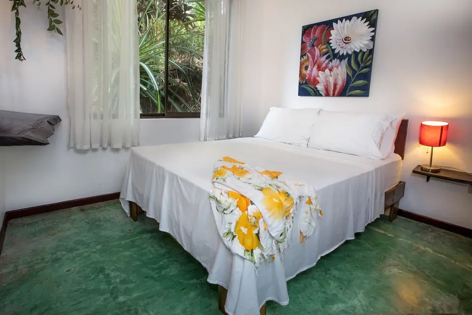 Villa Sol: a double bed with a yellow floral throw, a bedside lamp and a window onto green leaves