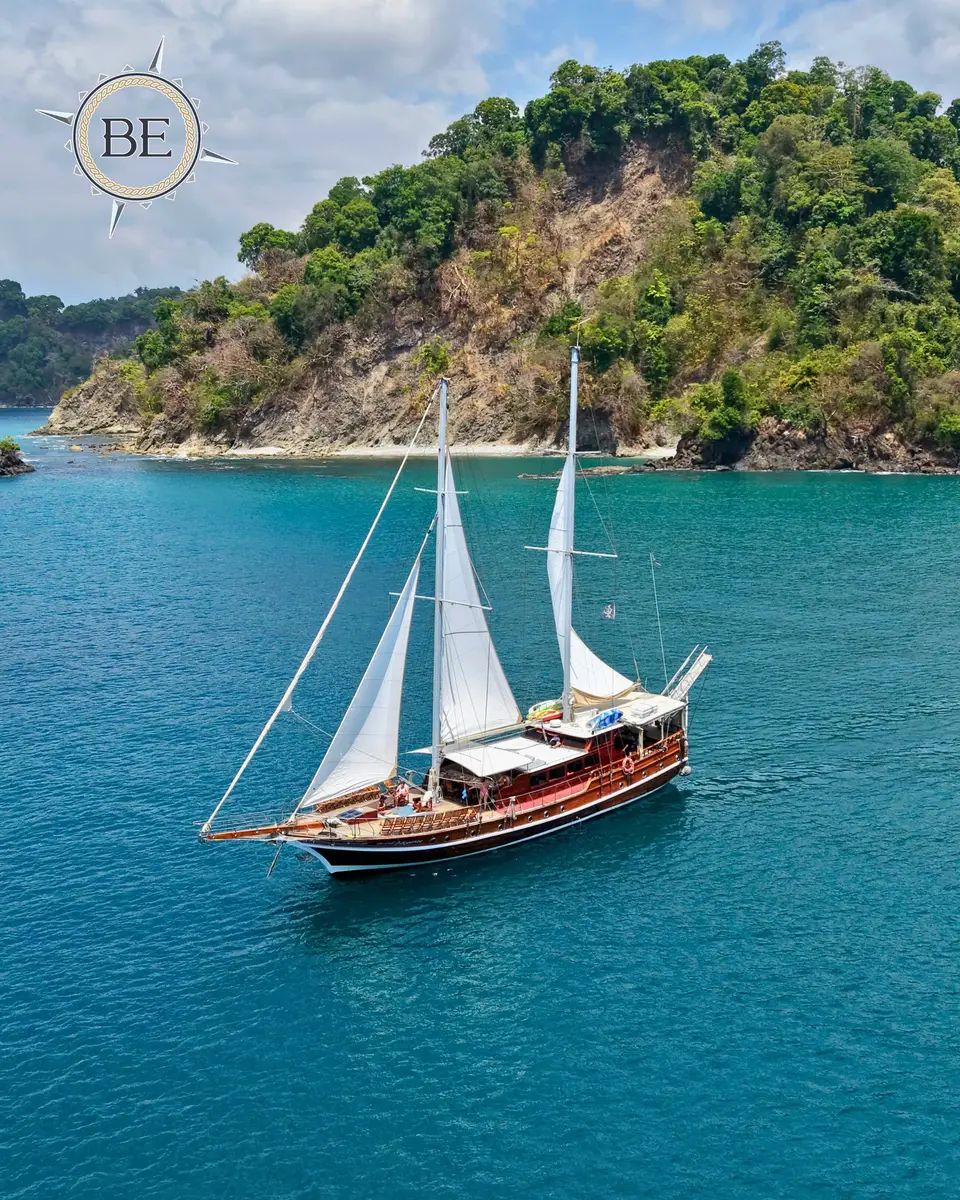 A two-masted wooden sailboat with white sails cruises past a rocky, forested island on turquoise water.