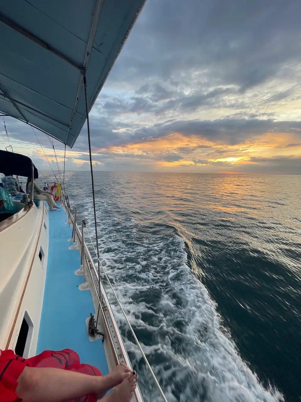 View along the side of a boat at sea at sunset, with white wake below and a golden sky over the horizon.