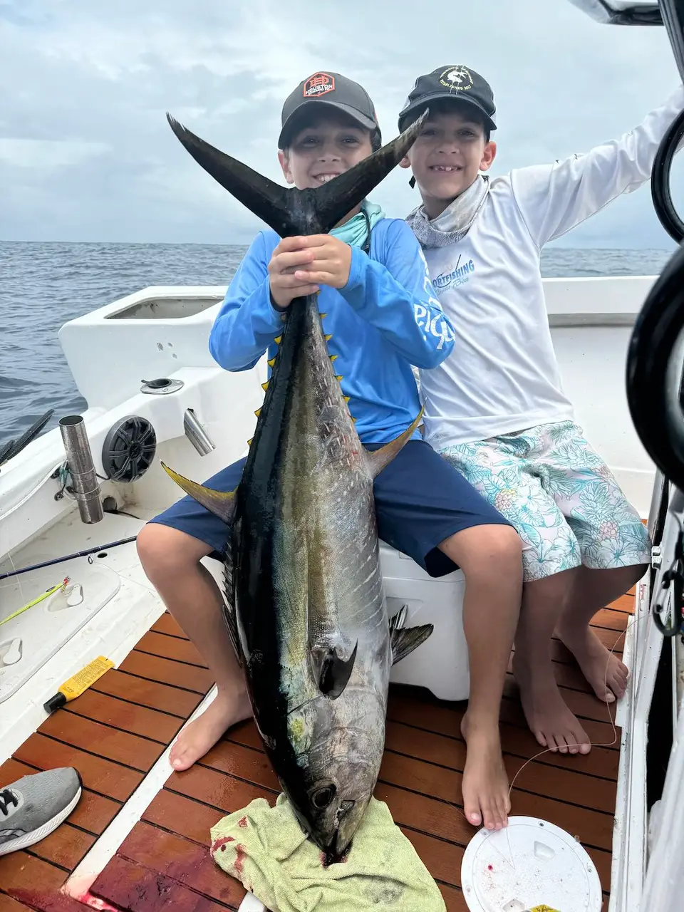Two boys in caps sit on a boat and hold up a big tuna between them.