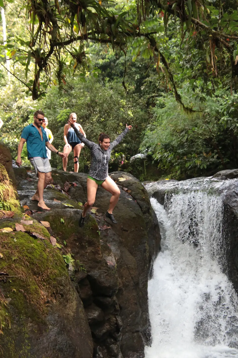 Three friends climb mossy boulders beside a small waterfall in the forest, one with her arms up in celebration.