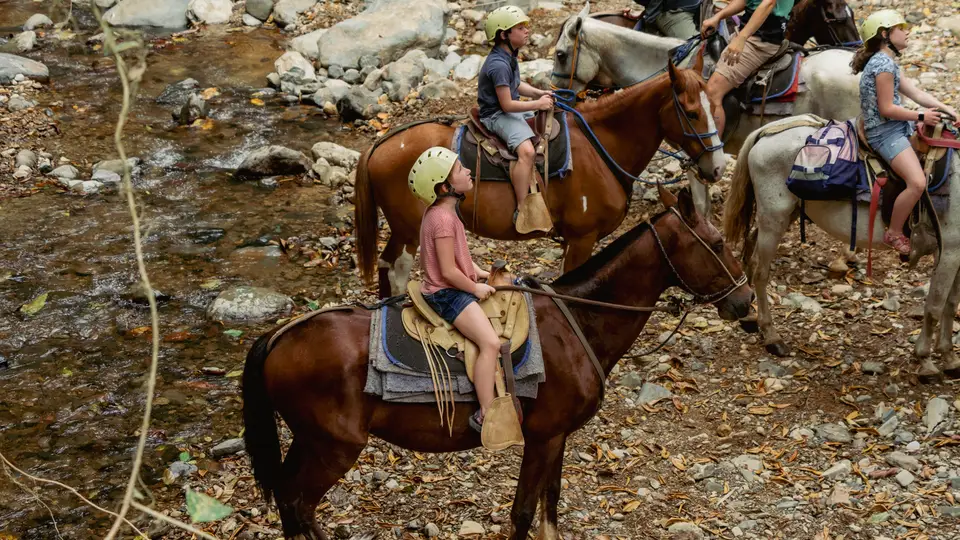 Children in helmets sit on horses at a rocky river crossing.