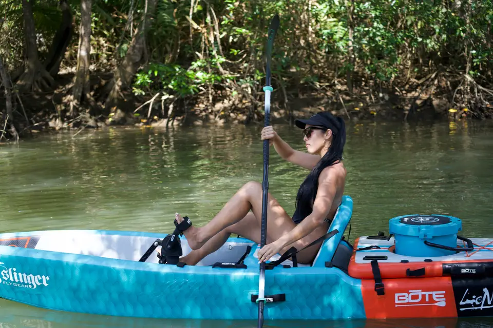 A woman in a cap and sunglasses paddles a sit-on kayak through a green mangrove channel.