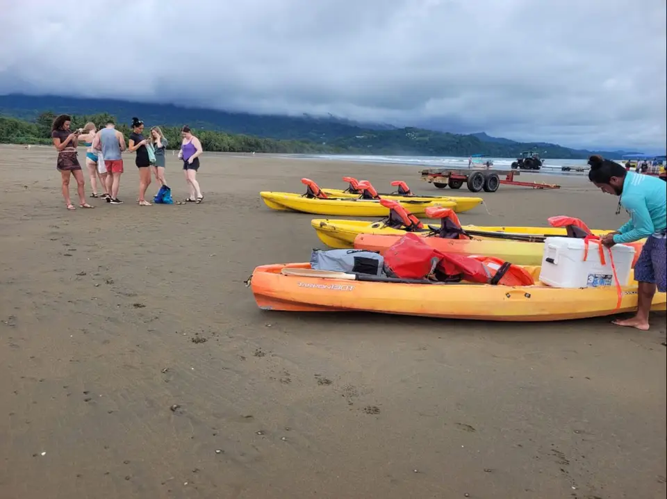 A guide loads a cooler onto orange and yellow kayaks on a long beach while the group waits under a cloudy sky.