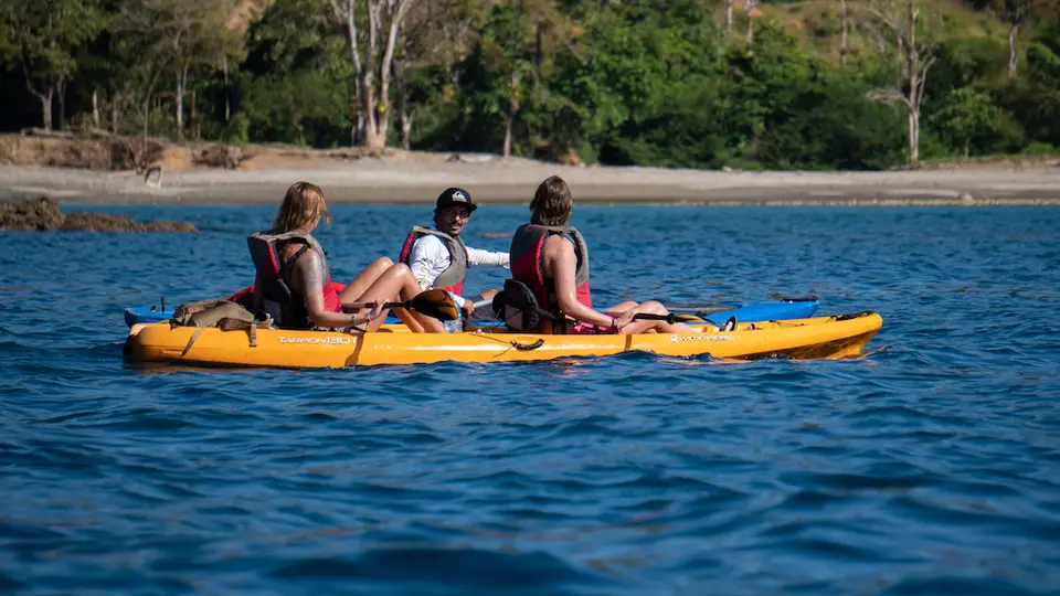 Tres personas con chaleco salvavidas en un kayak amarillo sobre agua azul intensa, con una playa boscosa detrás.