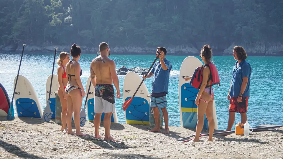 A group listens to an instructor on a pebbly beach beside upright paddleboards, turquoise water behind them.