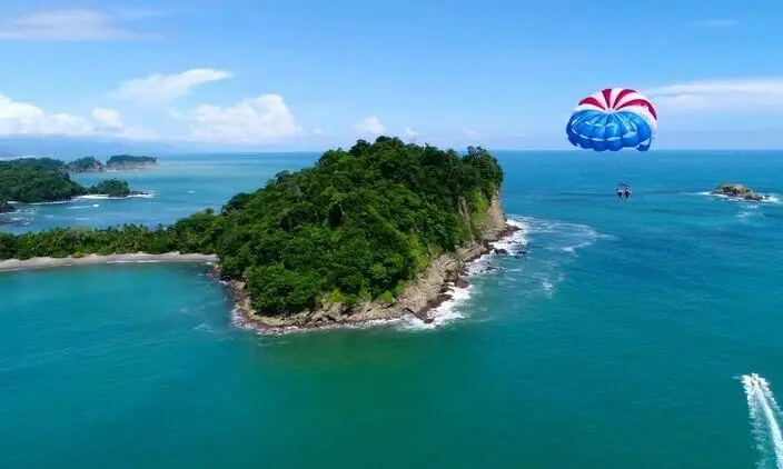 A parasail flies above turquoise water near a small forested island along the coast.