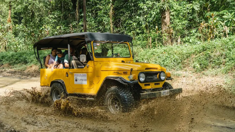 Smiling passengers ride in a yellow safari truck as it churns through mud on a jungle road.