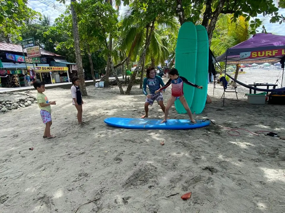 Children practice standing up on a blue surfboard on the sand under beachfront trees, with a surf tent behind.