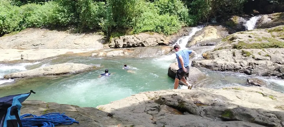 A guide stands on rocks beside a clear green river pool where two people swim below small cascades.