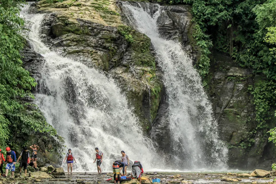 Rafters in life jackets wade at the foot of a tall twin waterfall in the rainforest.