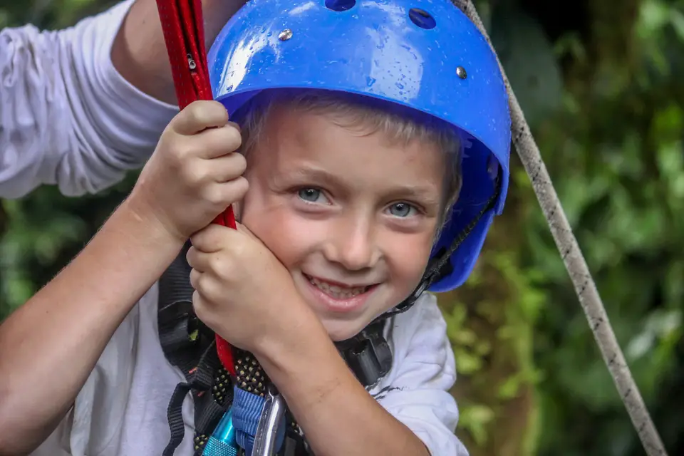 A smiling boy in a blue helmet grips a red rope while hanging in a harness.